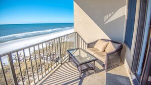 A balcony with a glass table and a cushioned seating area overlooking a beach.