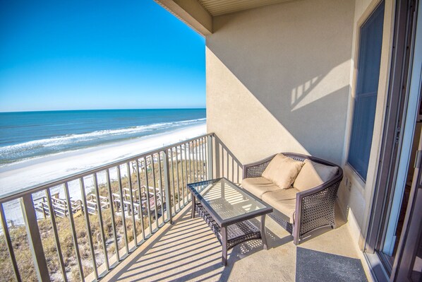 A balcony with a glass table and a cushioned seating area overlooking a beach.