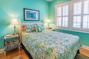 A bedroom with a bed, two bedside tables, a nightstand, a window with blinds, and a framed picture on the wall.