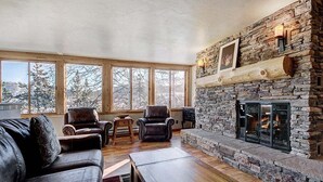A cozy living room with a stone fireplace, leather sofas, and large windows offering a view of trees.