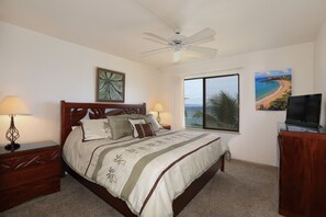 A bedroom with a bed, a ceiling fan, a nightstand, a desk with a TV, and a window with a beach view.