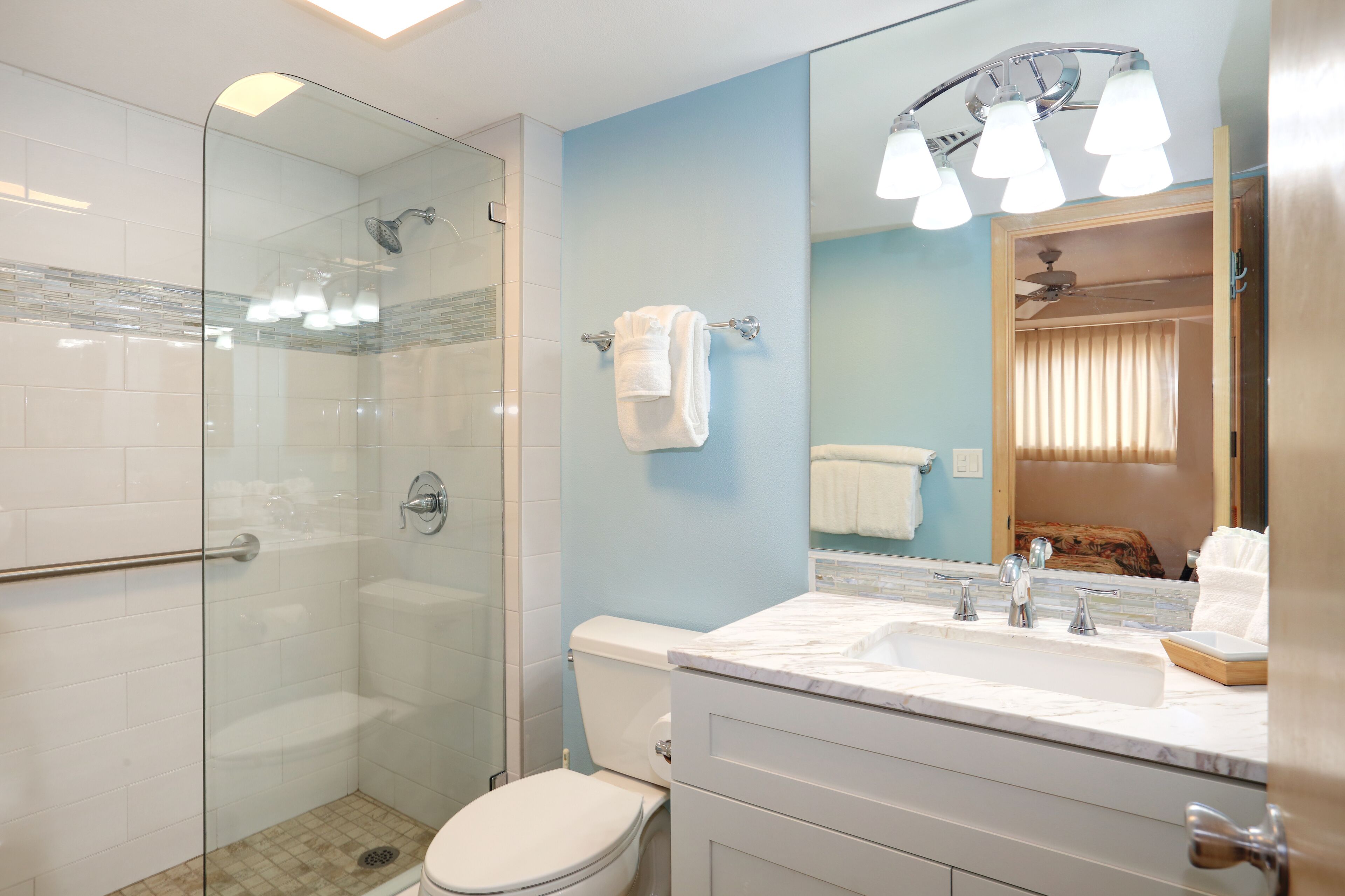 A modern bathroom with a glass shower enclosure, a white toilet, a marble sink, and a large mirror with overhead lighting.