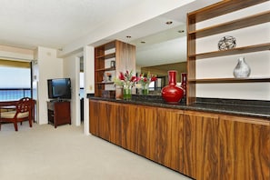 Ett hotellrum med en receptionsdisk i trä, en tv, en matplats med en stol och utsikt över havet.