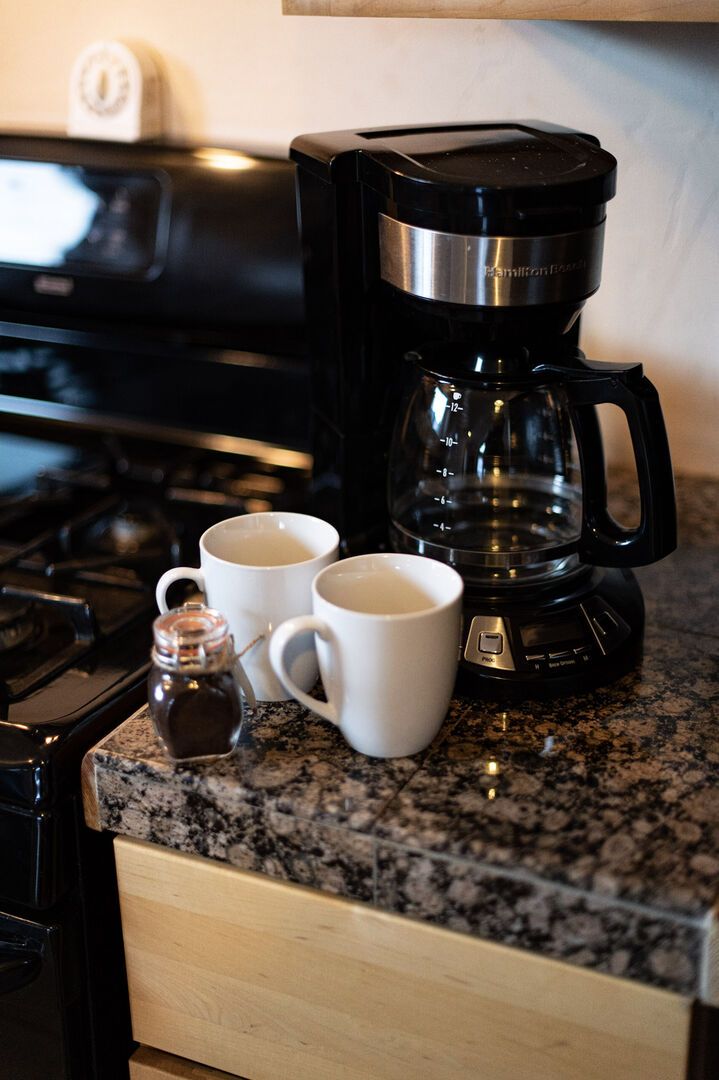Una encimera de cocina con una cafetera, dos tazas blancas y una botella pequeña de café.