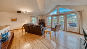 Un salón espacioso con un amplio sofá seccional marrón, una mesa de centro de madera y un mueble para la televisión. Grandes ventanales ofrecen vistas al exterior.