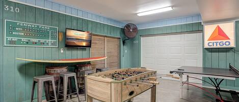 A room with a foosball table, a ping pong table, and a TV displaying a baseball game.