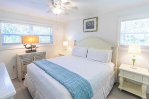 Une chambre à coucher avec un grand lit, deux tables de chevet avec des lampes, un ventilateur de plafond et une fenêtre donnant sur des arbres.