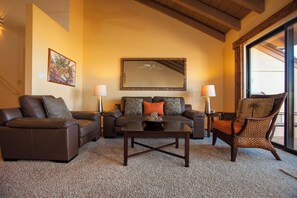 Un salon avec un canapé en cuir marron, une table basse en bois et un fauteuil en osier. On y trouve un ventilateur de plafond, un grand miroir et une fenêtre avec des rideaux.
