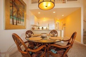 A dining area with a round table and six wicker chairs, a kitchen in the background, and a staircase leading to an upper level.