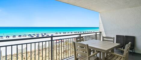 A balcony with a view of the beach and ocean, a table and chairs, and a railing.