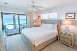 A hotel room with a bed, a nightstand, a lamp, a ceiling fan, a window with blinds, and a view of the ocean.