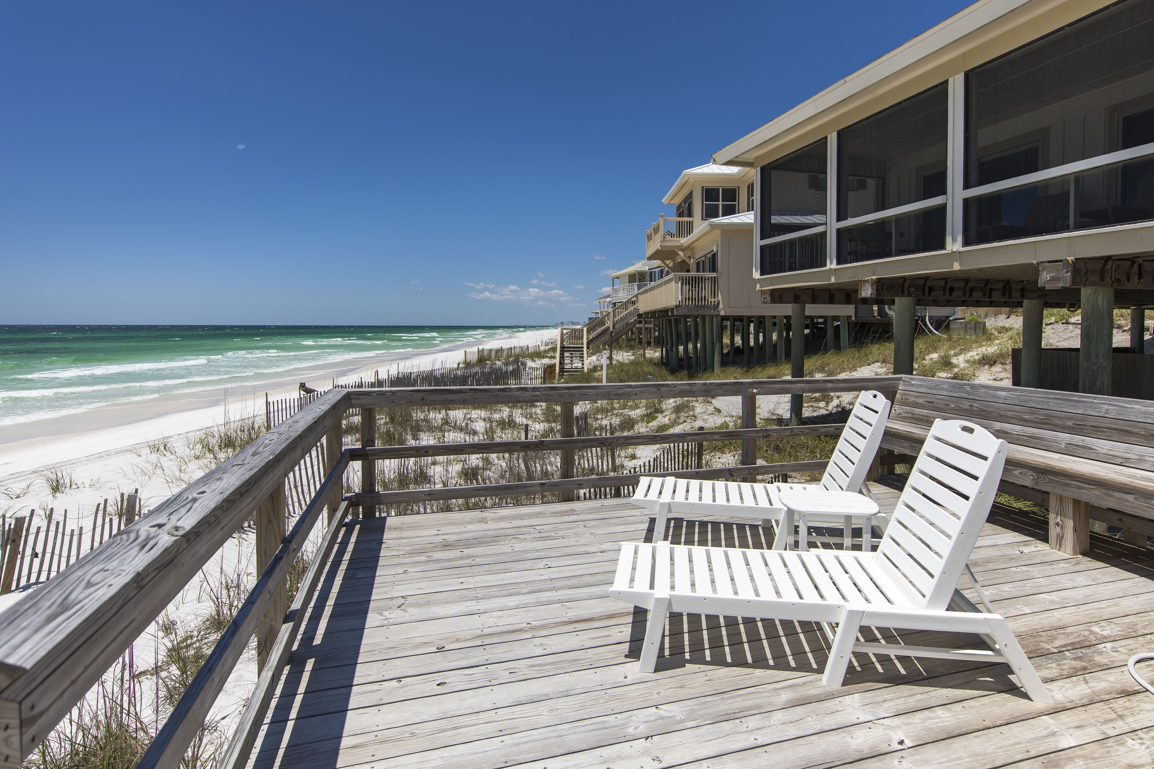A wooden deck with two white lounge chairs and a small table, overlooking a beach with a wooden fence and a building with large windows.