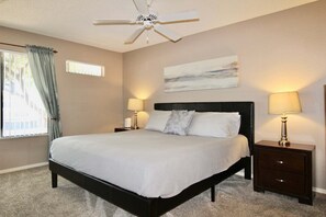 A bedroom with a bed, two bedside tables with lamps, a ceiling fan, a window with curtains, and a painting on the wall.
