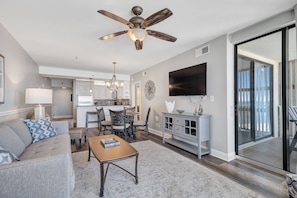 A modern living room with a ceiling fan, a flat-screen TV, a dining area, and a kitchen in the background.