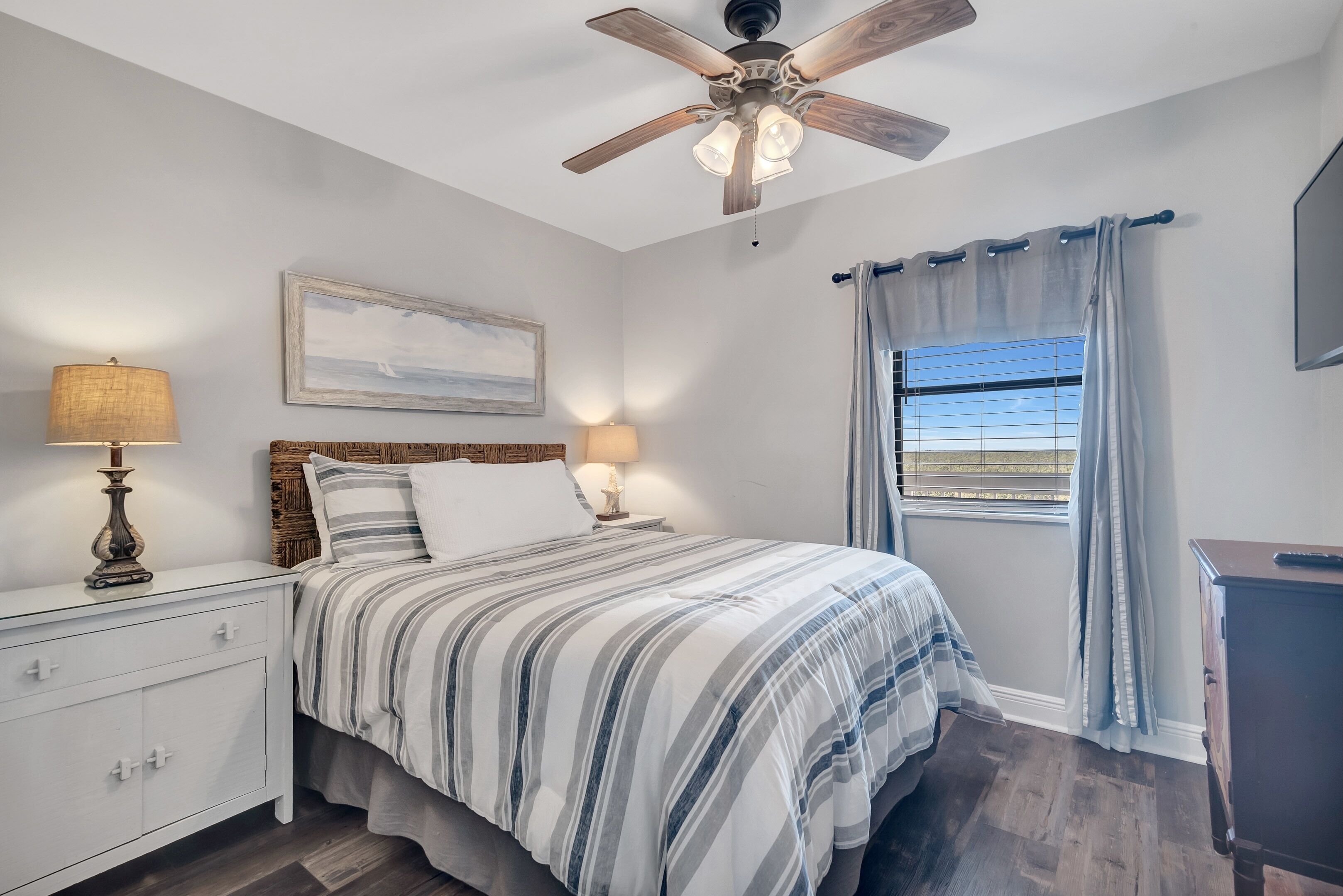 A bedroom with a bed, nightstands, a ceiling fan, a window with curtains, and a painting on the wall.