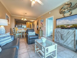 A spacious living room with a dining area, a kitchen in the background, and a large TV mounted on the wall. - Crystal Tower 407 - Beachfront pool, great amenities & private skywalk to beach! (Gulf Shores)
