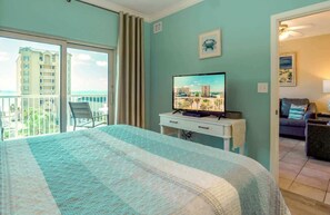 Habitación de hotel con una cama grande, un televisor sobre una mesa blanca, un balcón con una silla y vistas a un edificio.