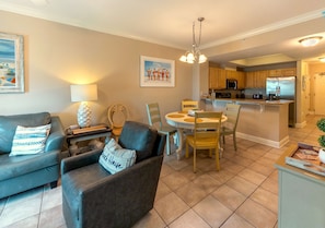 A living room with a sofa, armchair, and dining table with chairs. There is a kitchen area with cabinets and appliances. - Crystal Tower 407 - Beachfront pool, great amenities & private skywalk to beach! (Gulf Shores)