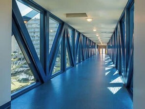 Un pasillo con ventanas amplias y suelo azul, que conduce a una puerta al final.