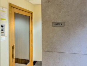 Una puerta de madera con un panel de vidrio esmerilado, un letrero que dice "SAUNA" sobre una pared texturizada, y un panel de control con varios botones e indicadores.