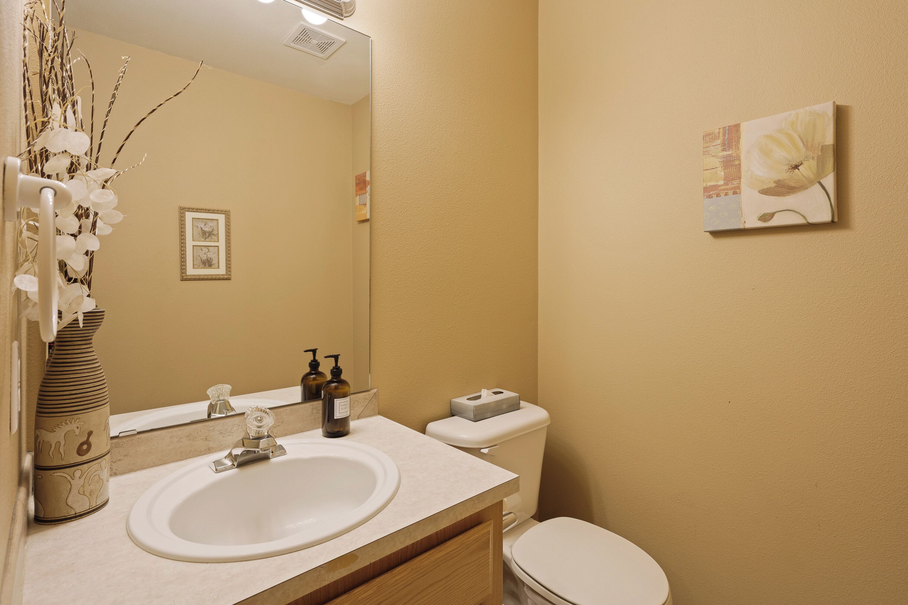 Een badkamer met een wit wastafel, spiegel en toilet. Links staat een vaas met takken en aan de muur hangt een lijst met een afbeelding.
