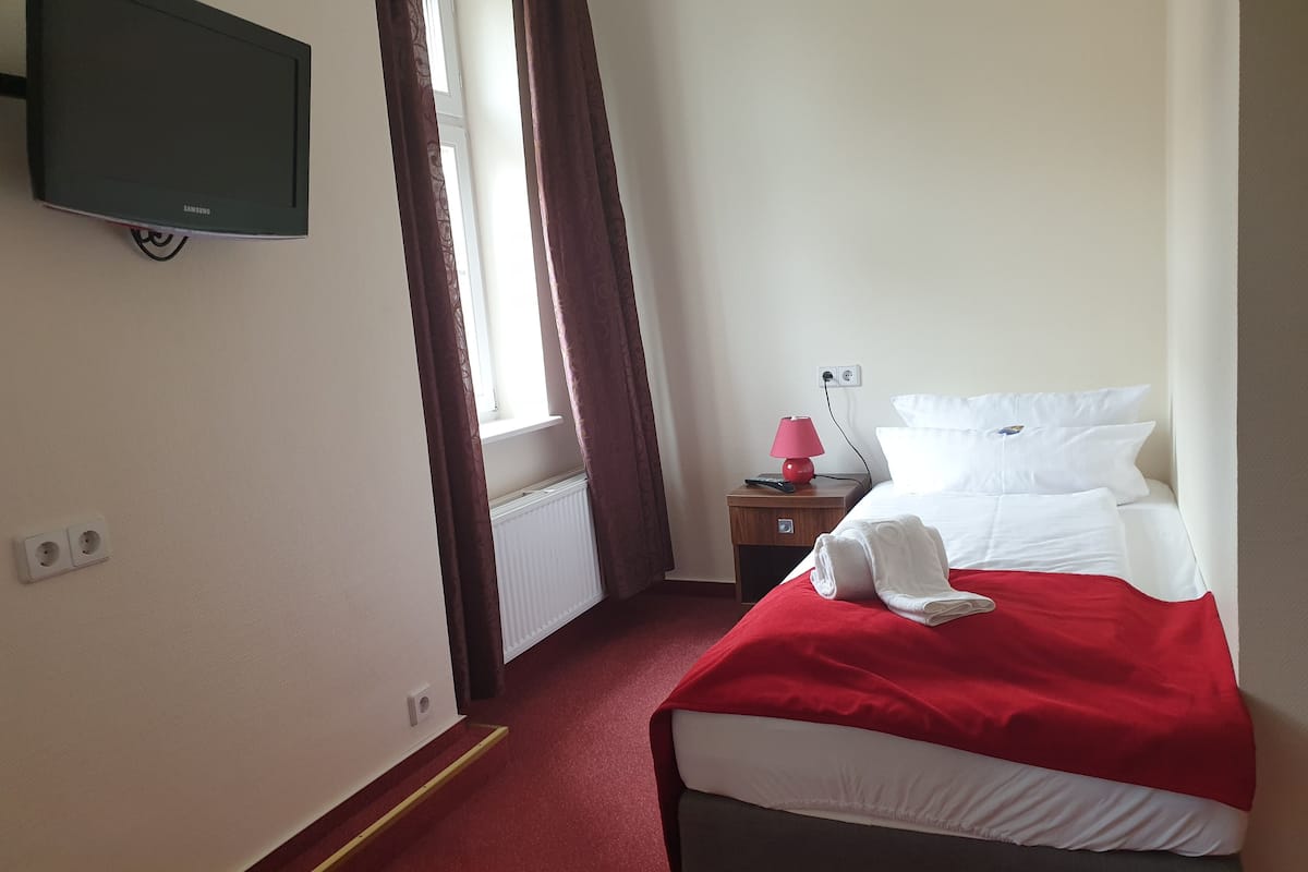 Une chambre d’hôtel avec un lit, un téléviseur fixé au mur, une table de chevet avec une lampe et une fenêtre avec des rideaux.