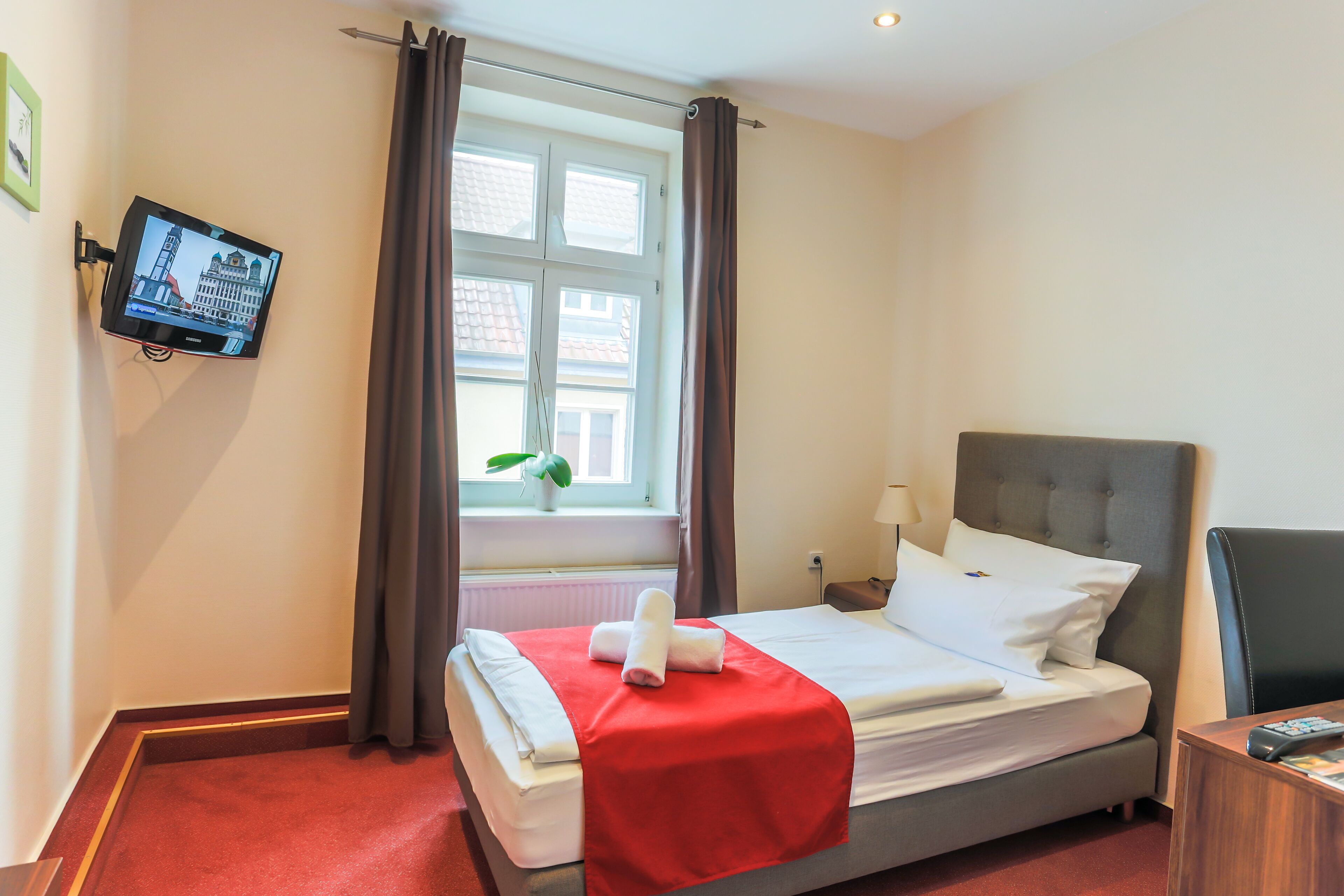 Habitación de hotel con cama, televisión montada en la pared, ventana con cortinas y planta en el alféizar.