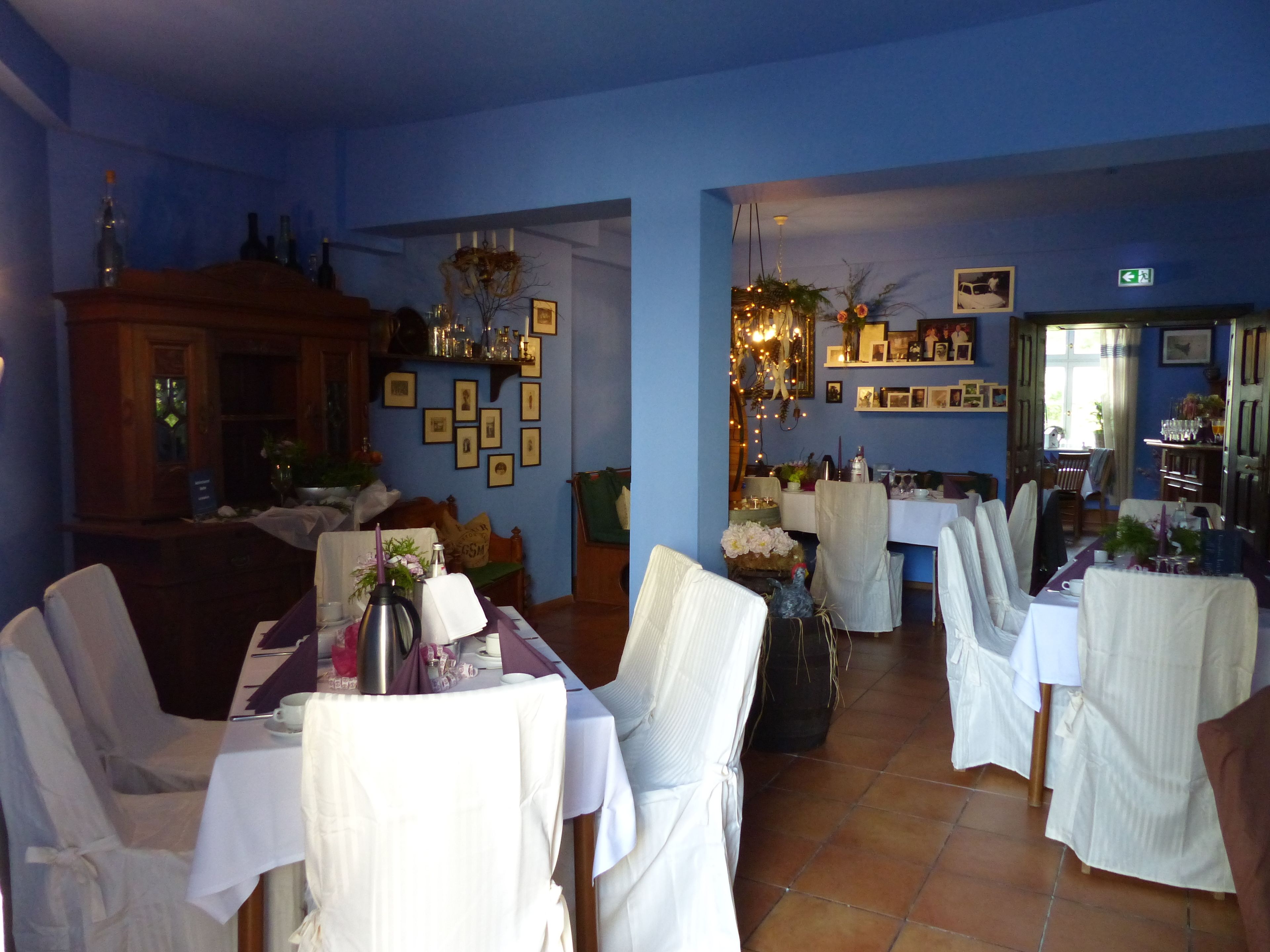A dining area with tables set for a meal, chairs, a chandelier, and a blue wall with framed pictures.