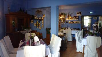 A dining area with tables set for a meal, chairs, a chandelier, and a blue wall with framed pictures.