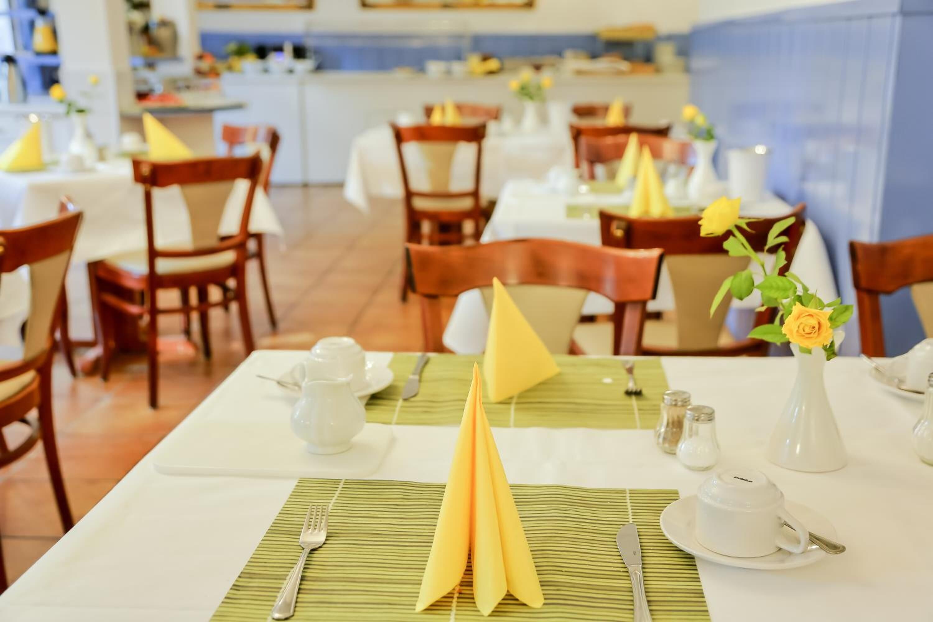 Un comedor con mesas dispuestas para dos personas, con servilletas dobladas con esmero, manteles blancos y un jarrón con una flor amarilla.