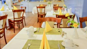 Un comedor con mesas dispuestas para dos personas, con servilletas dobladas con esmero, manteles blancos y un jarrón con una flor amarilla.