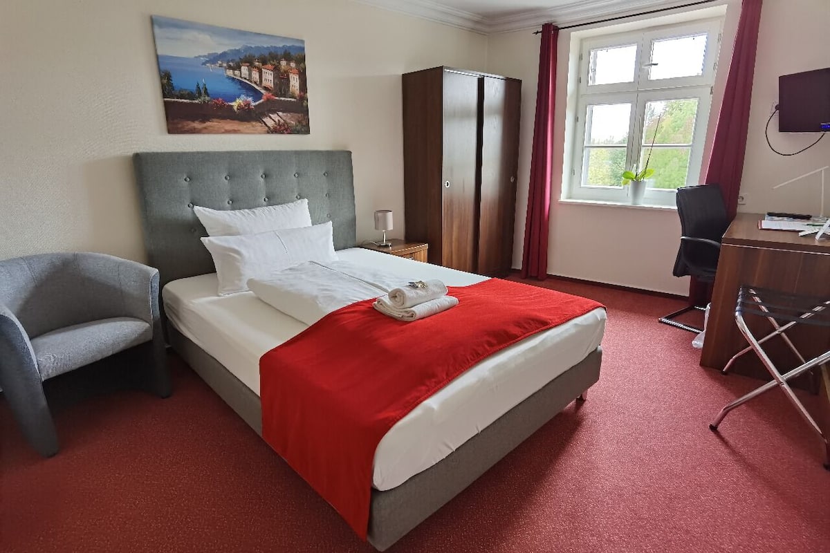 Une chambre d’hôtel avec un lit, une chaise, un bureau, une armoire, une télévision et une fenêtre avec des rideaux.