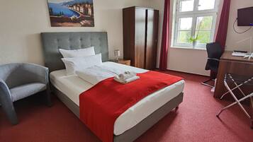 Ein Hotelzimmer mit Bett, Sessel, Schreibtisch, Kleiderschrank, Fernseher und einem Fenster mit Vorhängen.