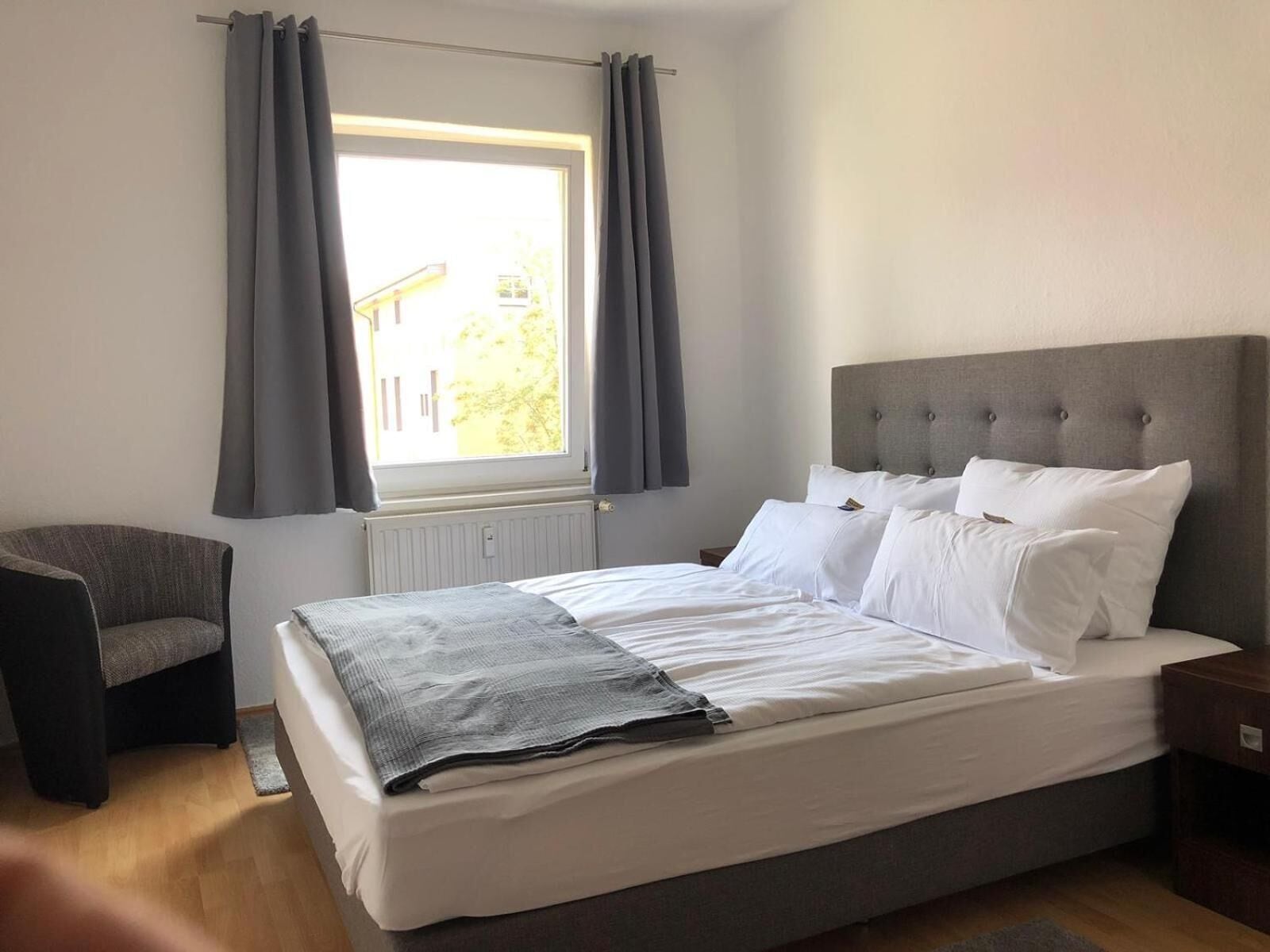 Ein Schlafzimmer mit Bett, Stuhl, einem Fenster mit Vorhängen und einem Heizkörper.