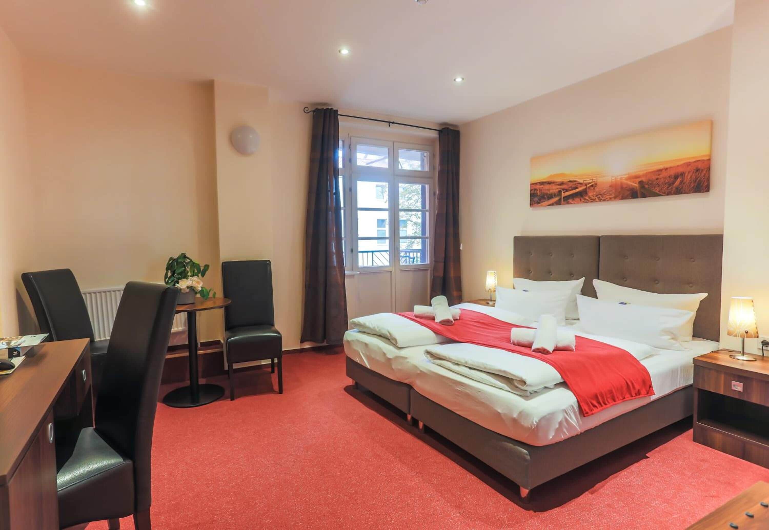 Una habitación de hotel con cama, escritorio, silla, ventana con cortinas y un cuadro en la pared.