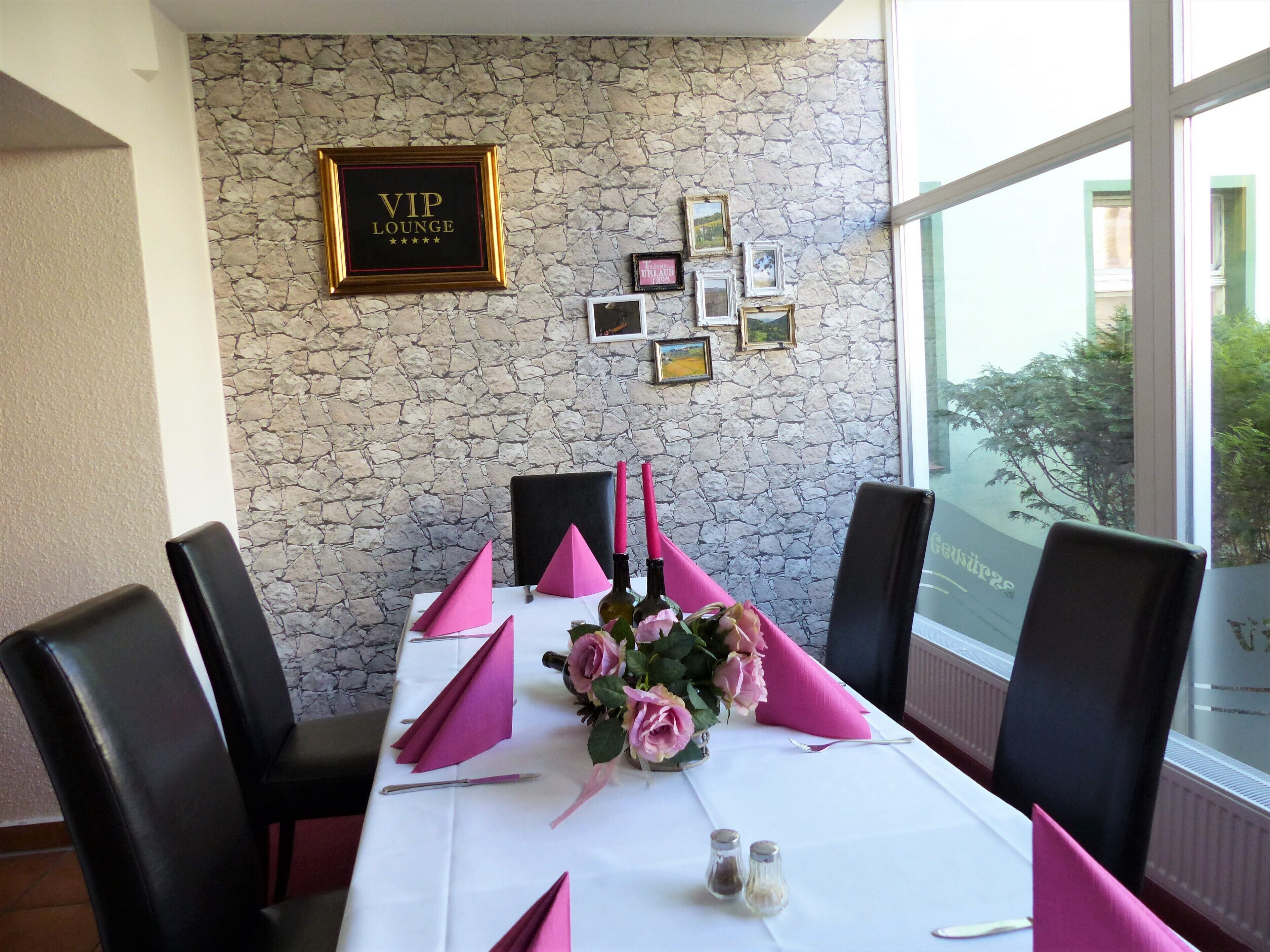 Un comedor con pared de piedra, un letrero que dice 'VIP Lounge', cuadros enmarcados, servilletas rosas y un jarrón con flores.