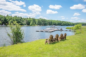 Une vue sur le lac avec des bateaux amarrés, une zone herbeuse avec des chaises en bois, et une colline boisée en arrière-plan.