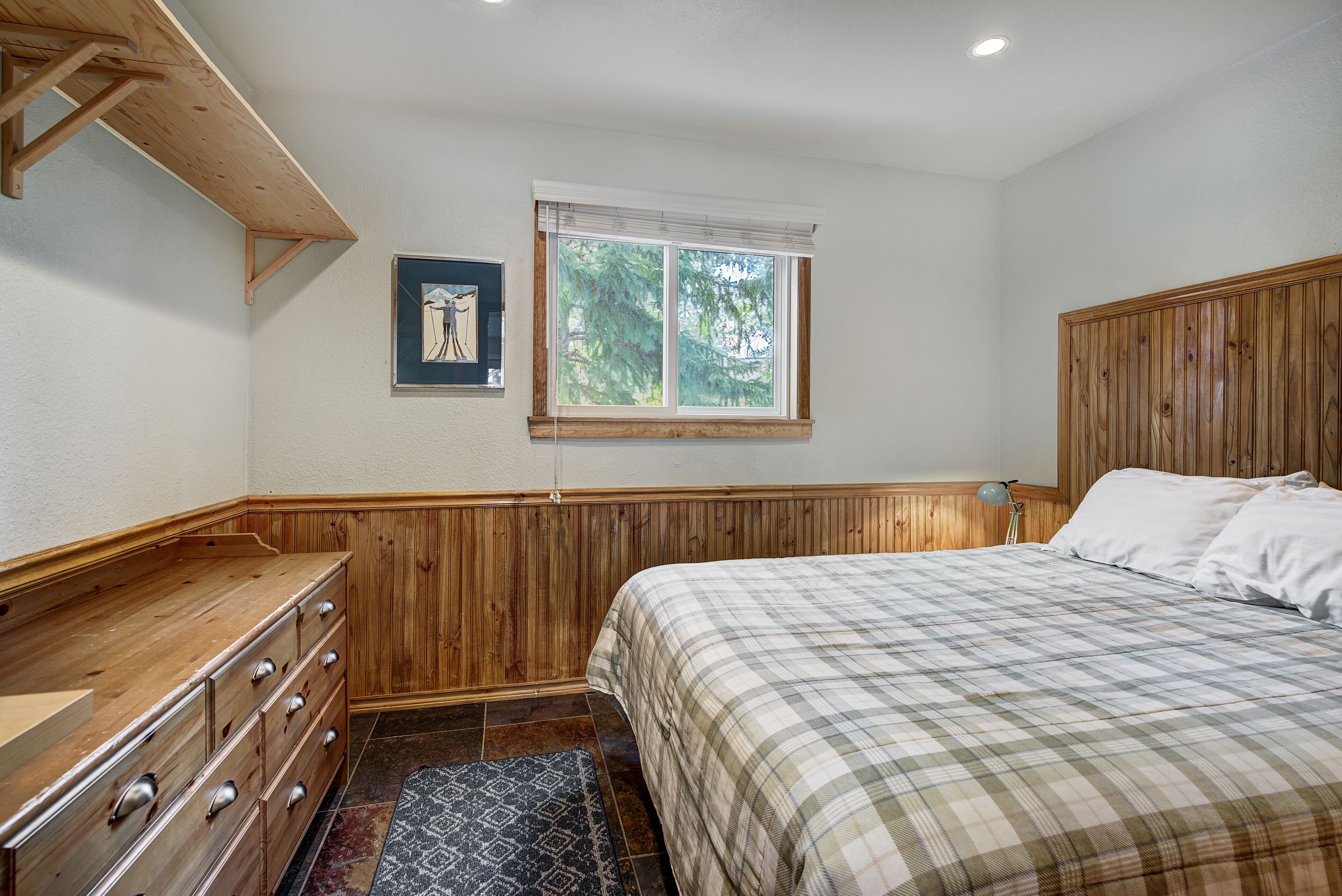 A bedroom with a wooden headboard, a bed with a plaid bedspread, a dresser with drawers, a window with blinds, and a framed picture on the wall.