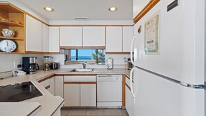 A kitchen with white cabinets, a dishwasher, a coffee maker, and a window with a view. - Experience Unforgettable Ocean Views at the Charming Kihei Beach #407 (Kihei)