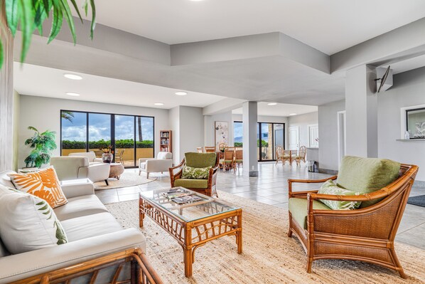 Un amplio salón con una gran mesa de centro de vidrio, muebles de mimbre y vistas al exterior.