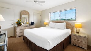 A hotel room with a large bed, bedside lamps, a ceiling fan, a dresser, a mirror, and a window with a view. - Experience Unforgettable Ocean Views at the Charming Kihei Beach #407 (Kihei)