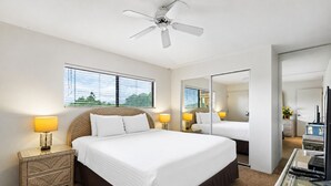 Ein Hotelzimmer mit einem großen Bett, zwei Nachttischlampen, einem Deckenventilator, einem großen Fenster mit Jalousien, einem Spiegel und einem Schreibtisch mit Computer.