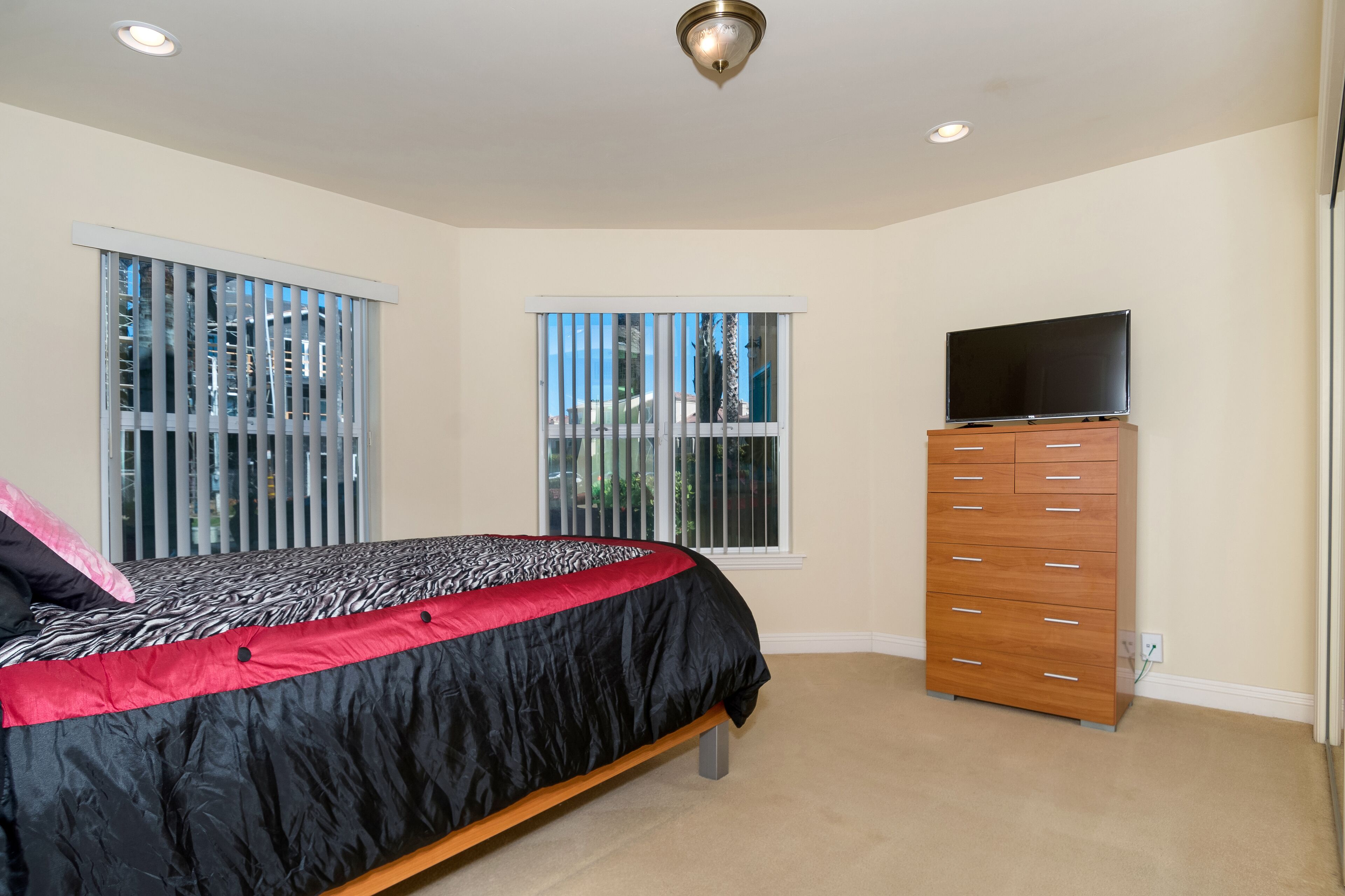 Quarto com cama, penteadeira, televisão e duas janelas com persianas.