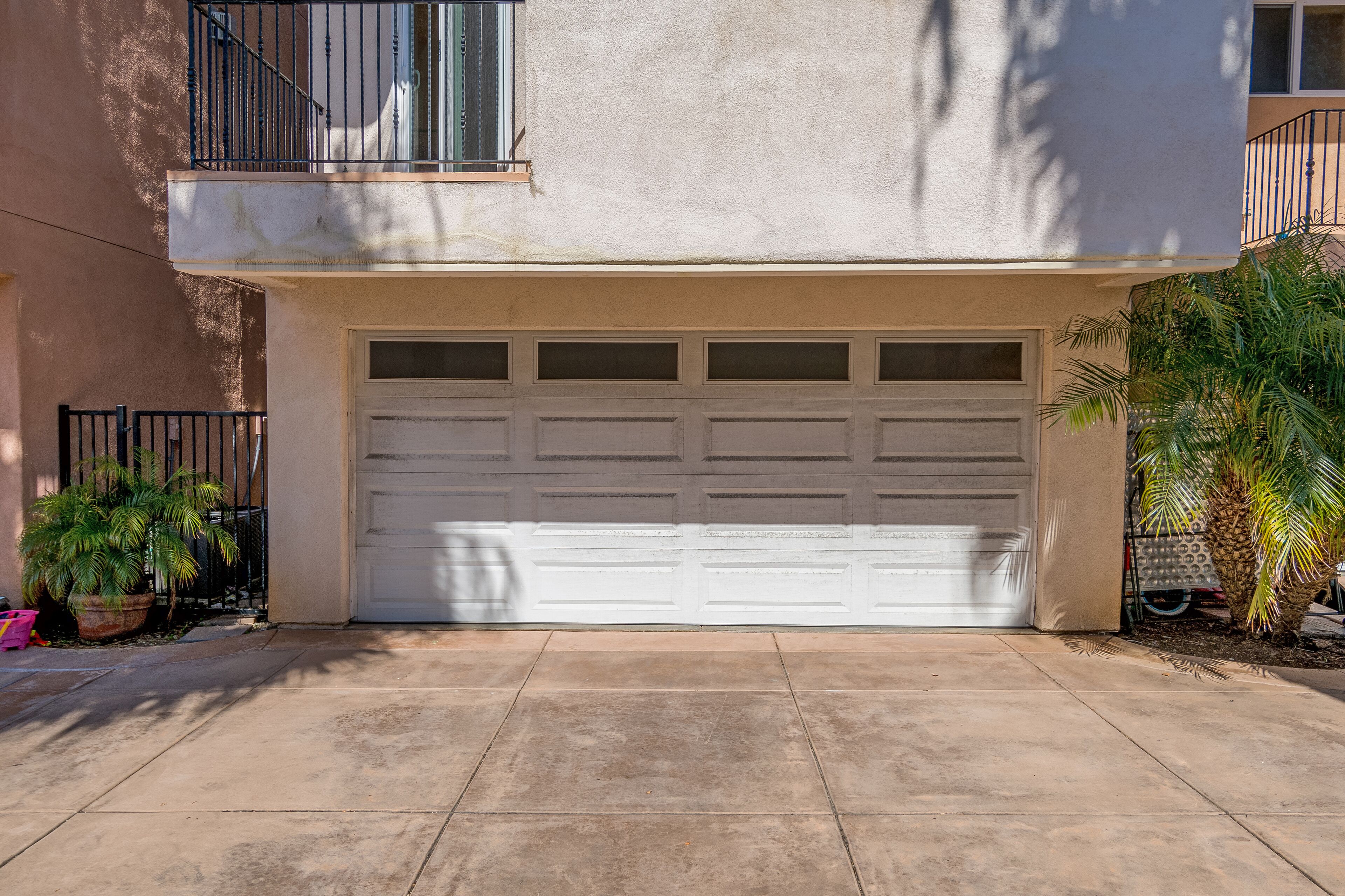 A residential garage with a white door, a balcony with black railings, and a potted plant to the left.