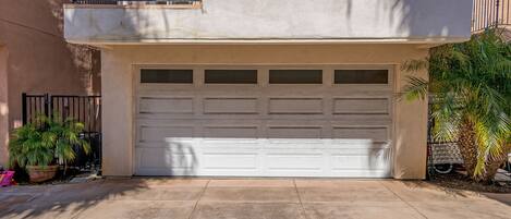 A residential garage with a white door, a balcony with black railings, and a potted plant to the left.