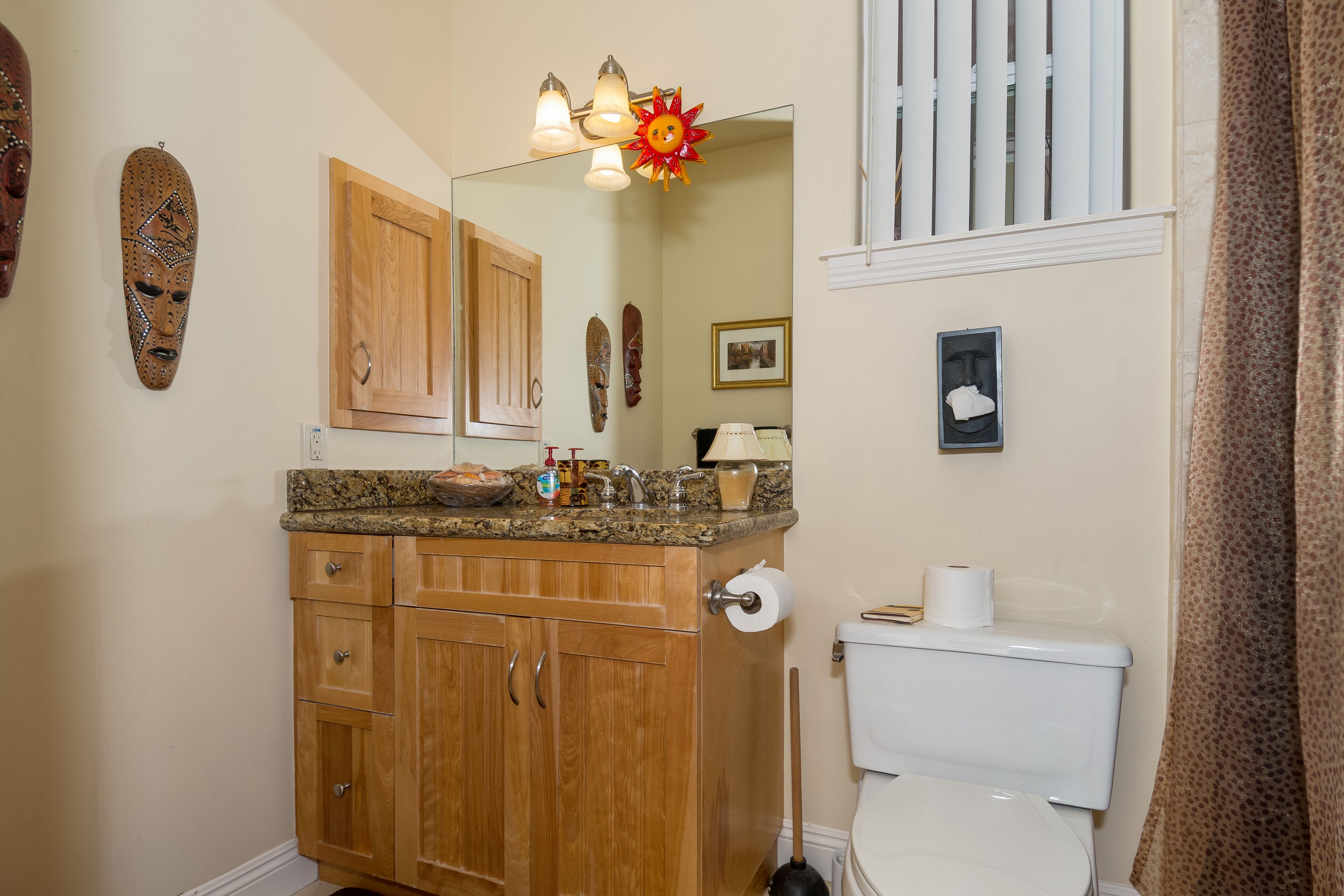 A bathroom with wooden cabinets, a granite countertop, a toilet, a mirror, and a sun-shaped light fixture.