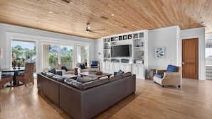 Een ruime woonkamer met een grote hoekbank, een eethoek met rieten stoelen en een televisie op een witte tv-meubel.