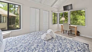 Un dormitorio con una cama grande, un escritorio y un ventilador de techo. Varias ventanas ofrecen luz natural y vistas al exterior.