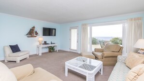 A living room with a beige sofa, a white wicker coffee table, a beige armchair, a white side table with a lamp, and a television mounted on the wall. - Gulf front townhouse with community pool, incredible views- Blue Mountain Beach (Santa Rosa Beach)