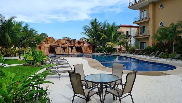 A pool area with lounge chairs and a table, surrounded by palm trees and a building with balconies. - Sunrise 30, beautiful Condo In the Heart of Town (Tamarindo)